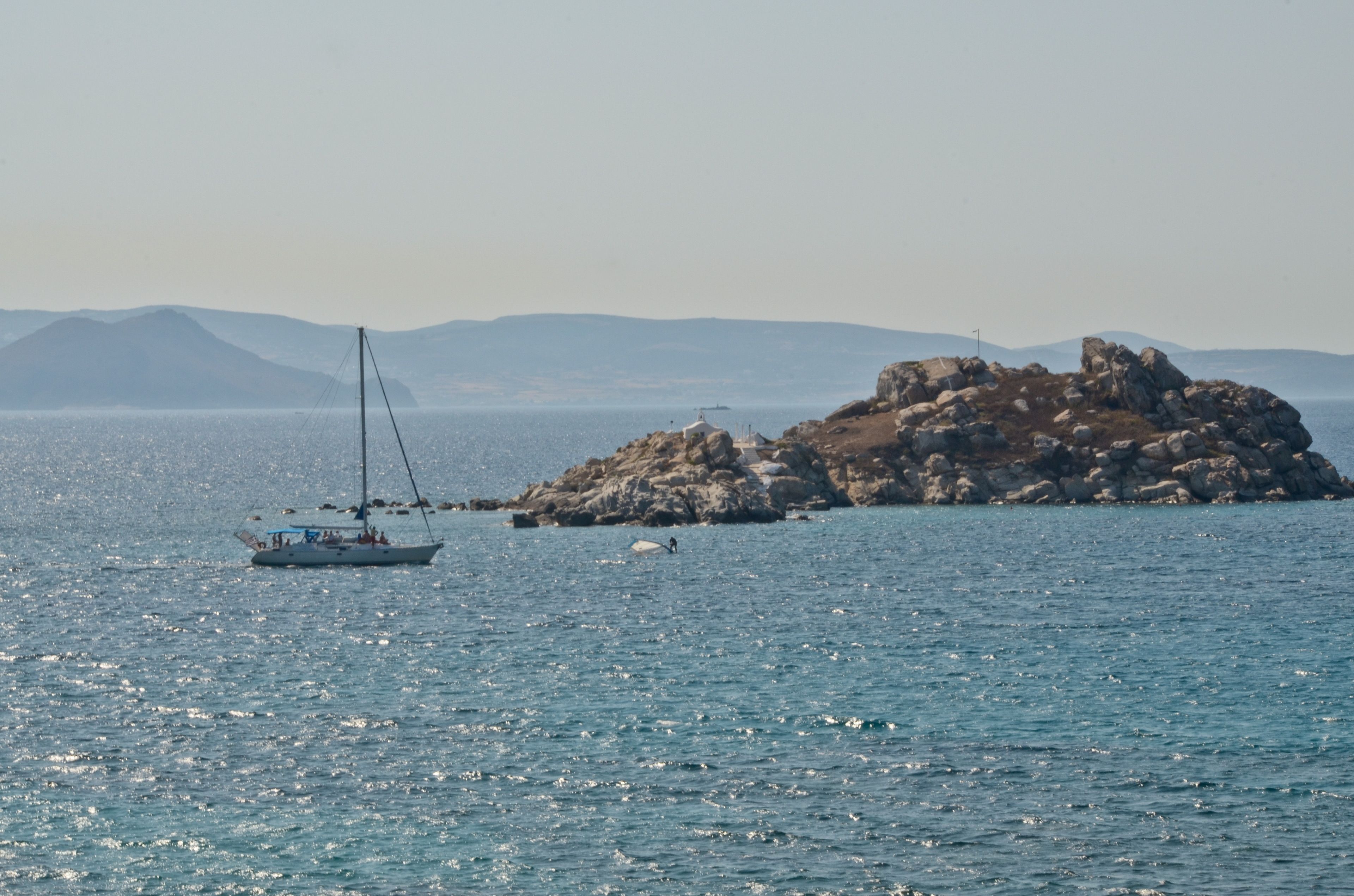 Un voilier navigue près d’une île rocheuse, sur une eau calme, avec des montagnes qui se dessinent à l’horizon.
