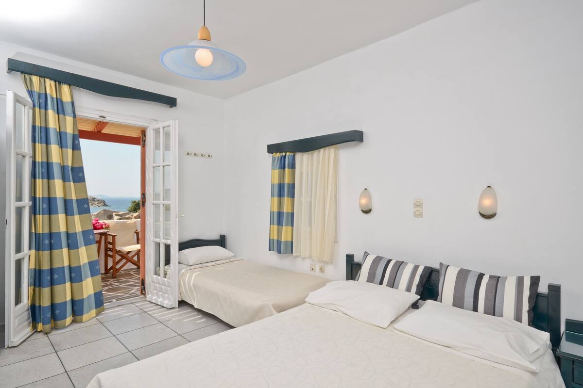 Ein Hotelzimmer mit zwei Betten, Blick auf das Meer durch eine offene Tür und einem kleinen Balkon mit Tisch und Stühlen.