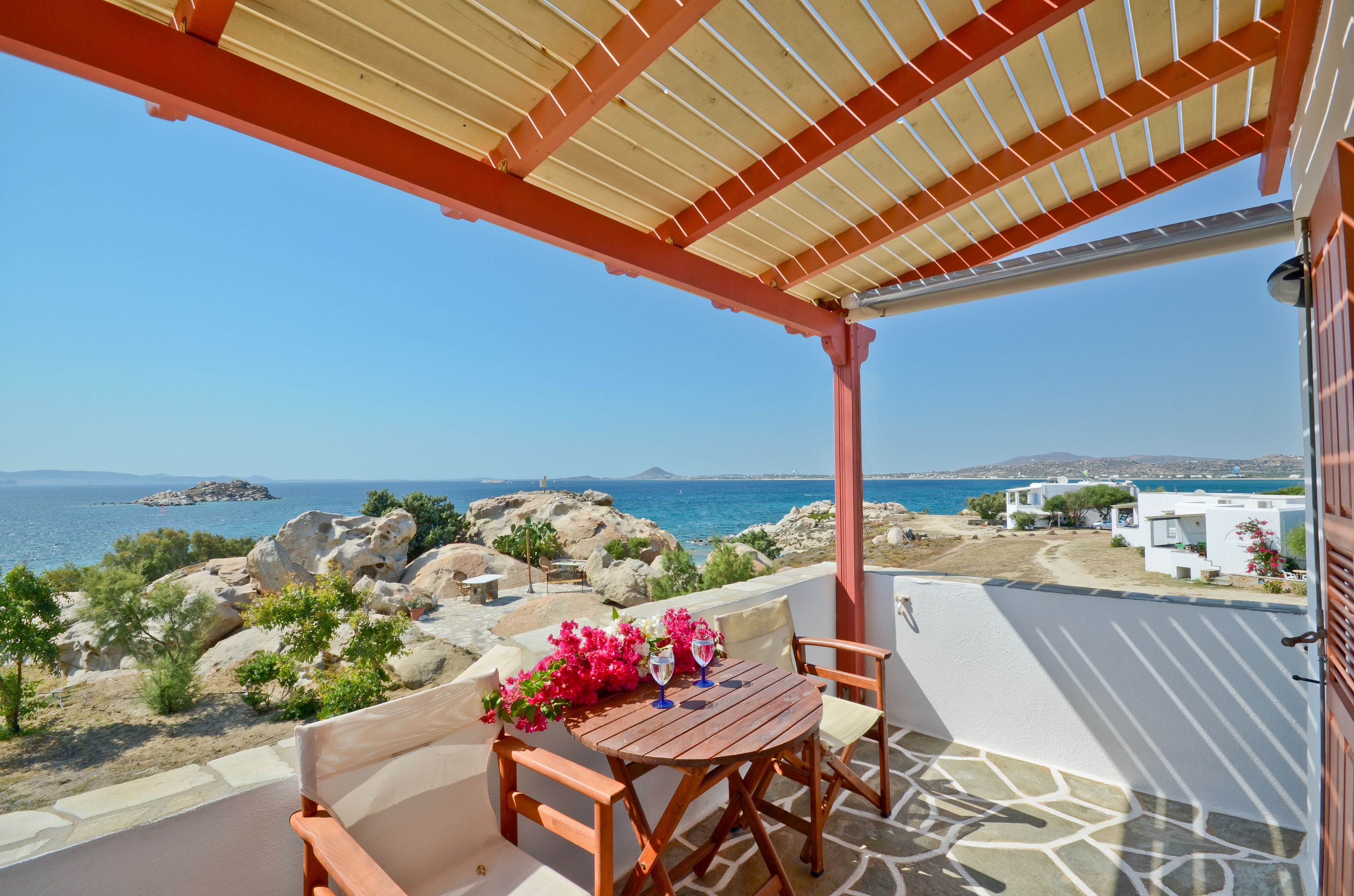 A wooden table with chairs and a tablecloth, set on a balcony with a view of the sea and rocky landscape.