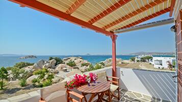 A wooden table with chairs and a tablecloth, set on a balcony with a view of the sea and rocky landscape.