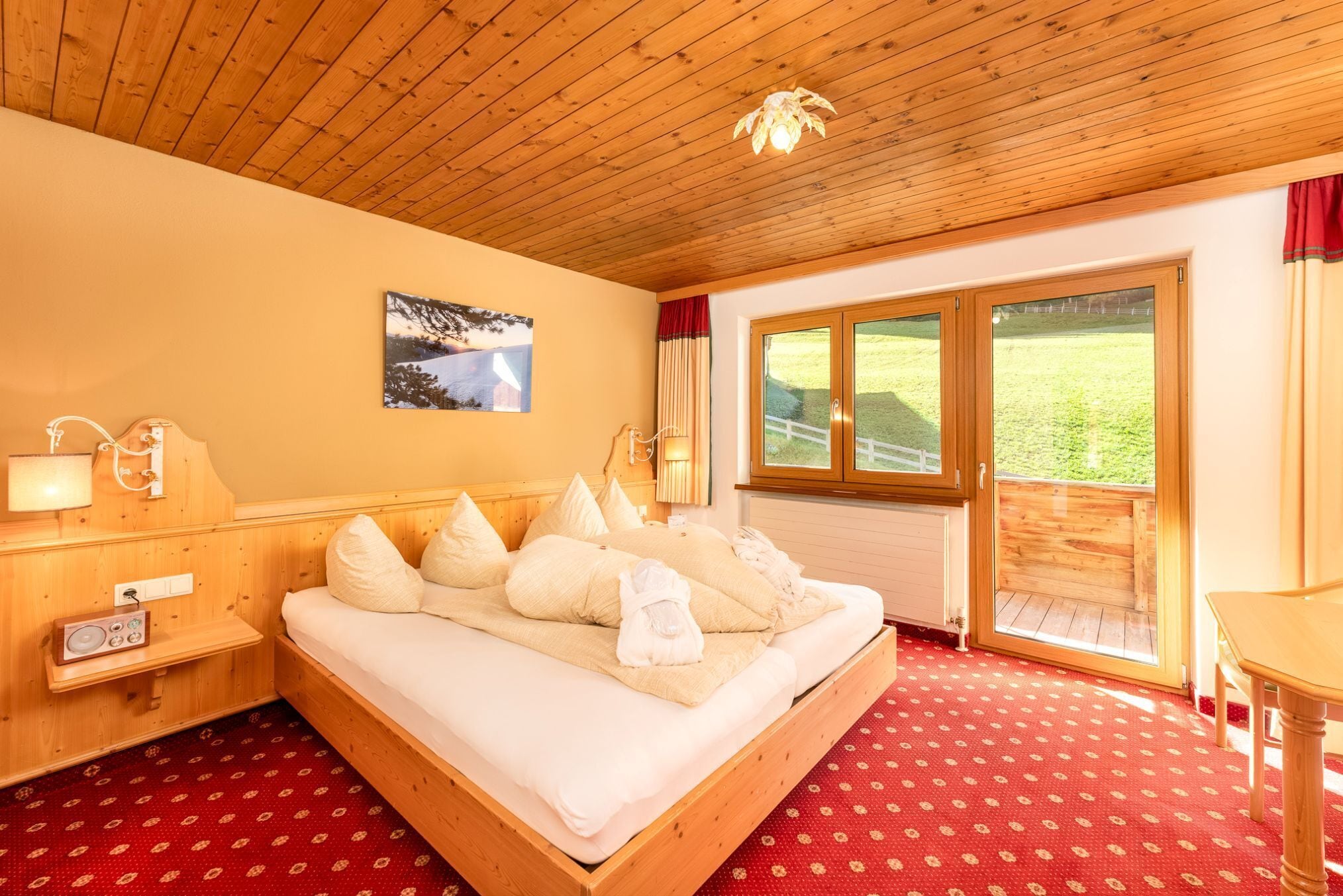 A wooden-framed bed with white linens and pillows, a wooden nightstand with a radio, a picture on the wall, and a large window with a view of greenery.