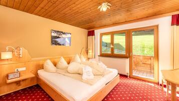 A wooden-framed bed with white linens and pillows, a wooden nightstand with a radio, a picture on the wall, and a large window with a view of greenery.
