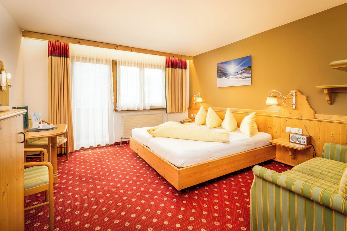 Ein Hotelzimmer mit einem hölzernen Bett, einem Schreibtisch mit Stuhl, einem Radio und einem Gemälde an der Wand.