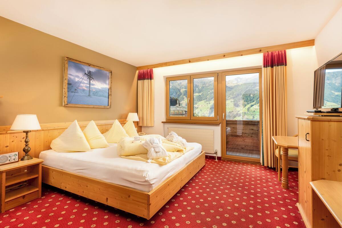 Ein Hotelzimmer mit einem hölzernen Bett, einem Bild an der Wand, einem Fenster mit Vorhängen und einem Fernseher auf einer Holzkommode.