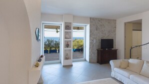 Un salon avec un canapé blanc, un téléviseur posé sur un meuble en bois et une vue sur la mer par une porte vitrée.