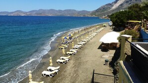 A beach with rows of sun loungers and umbrellas, a clear blue sea, and mountains in the background. - Hotel Plaza (Kalymnos)
