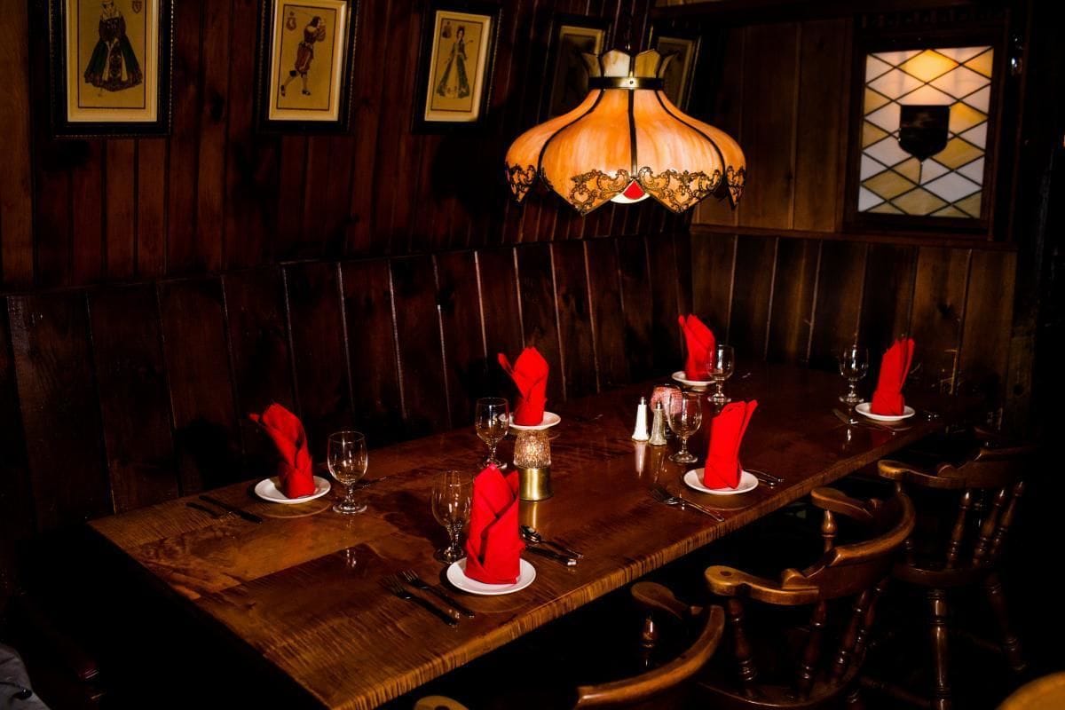 A long wooden dining table with red napkins, glasses, and cutlery, set in a dimly lit room with wooden paneling and framed pictures on the walls.