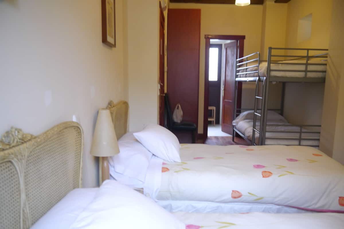 A room with two single beds, a wooden headboard, a bedside lamp, and a bunk bed in the background.
