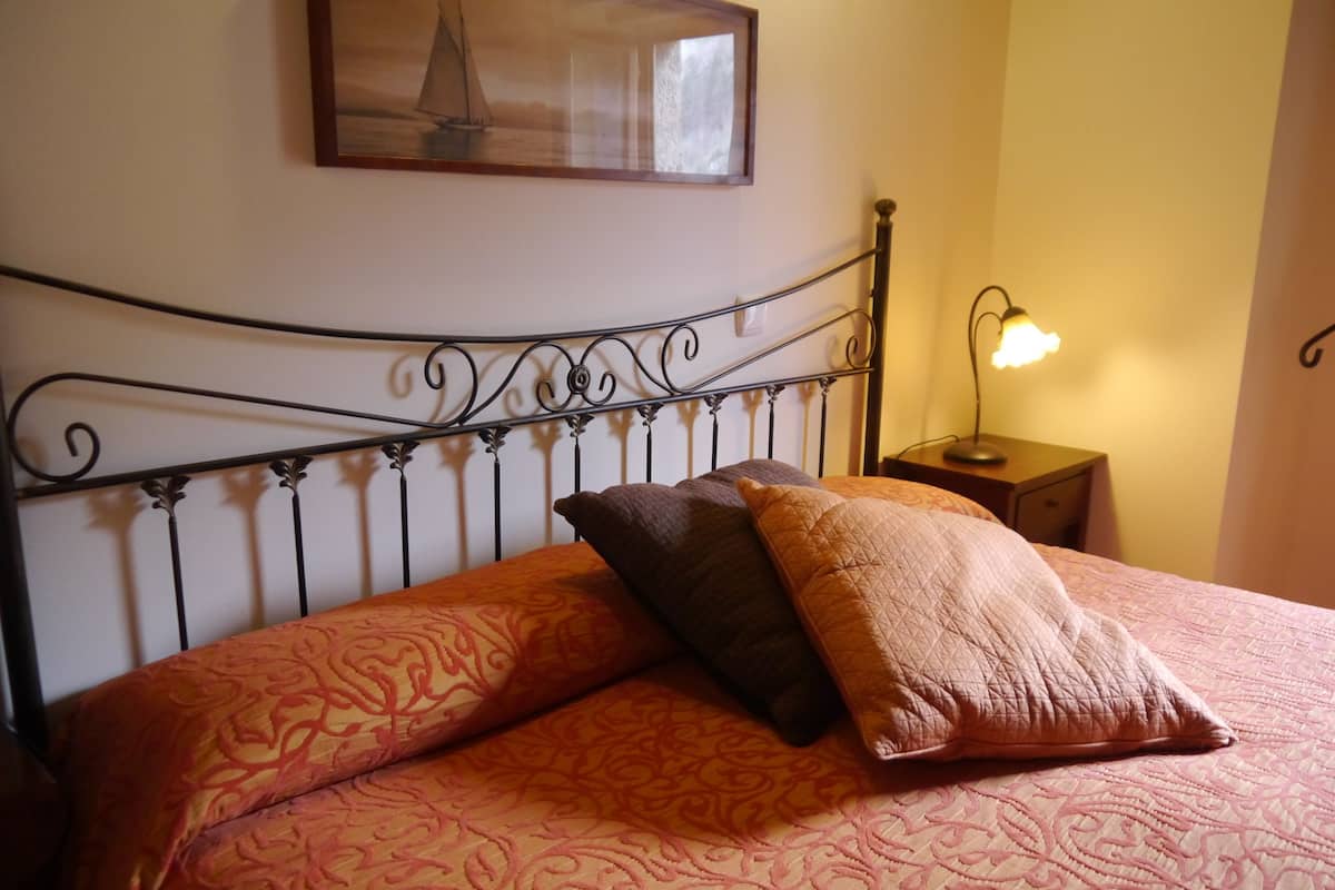A bed with a patterned bedspread and two pillows, a wall-mounted picture, a bedside lamp, and a nightstand.