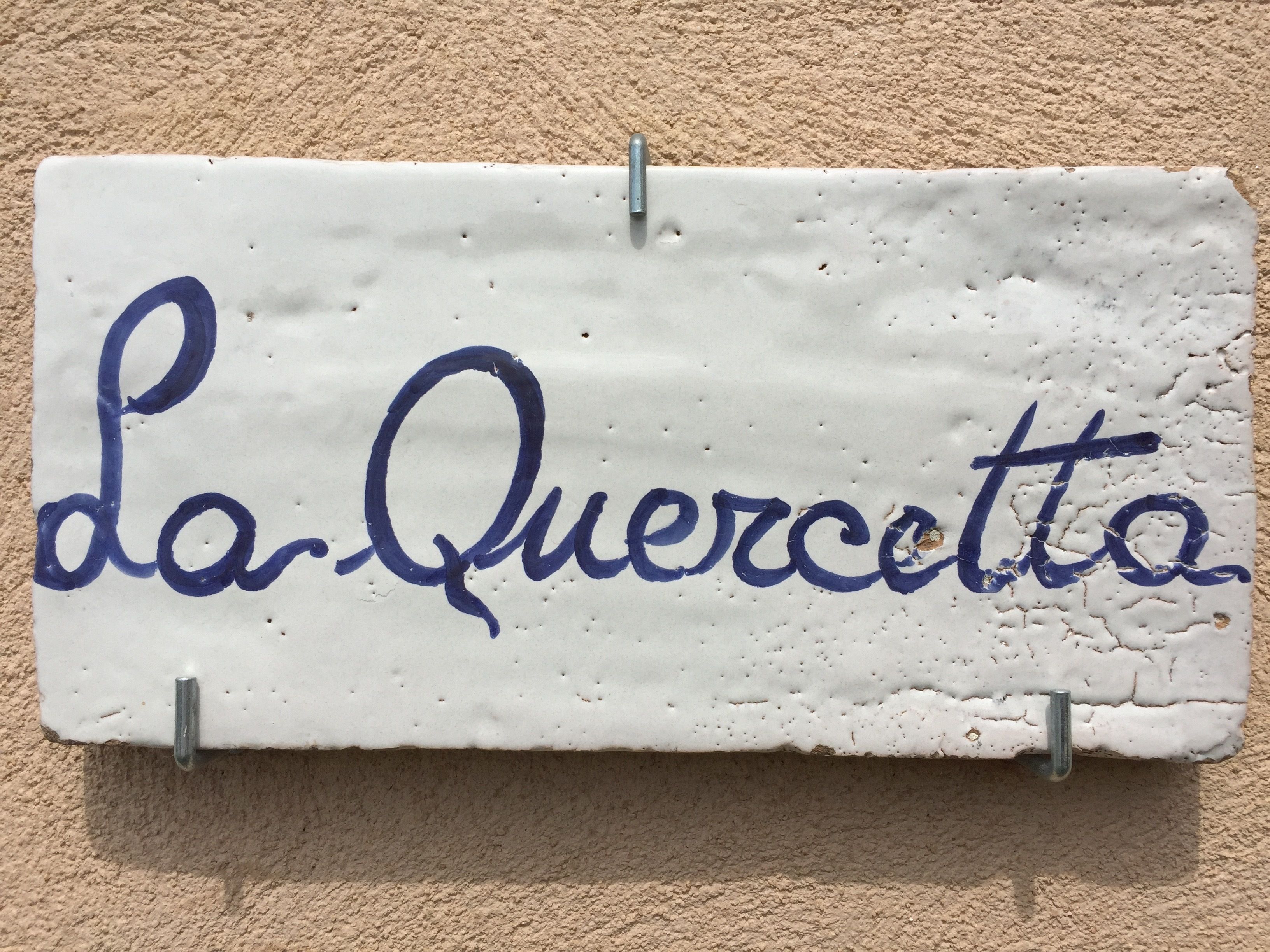 Ein weißes Keramikschild mit blauer verschnörkelter Schrift, die "La Queratta" zeigt, an einer strukturierten beige farbenen Wand befestigt.