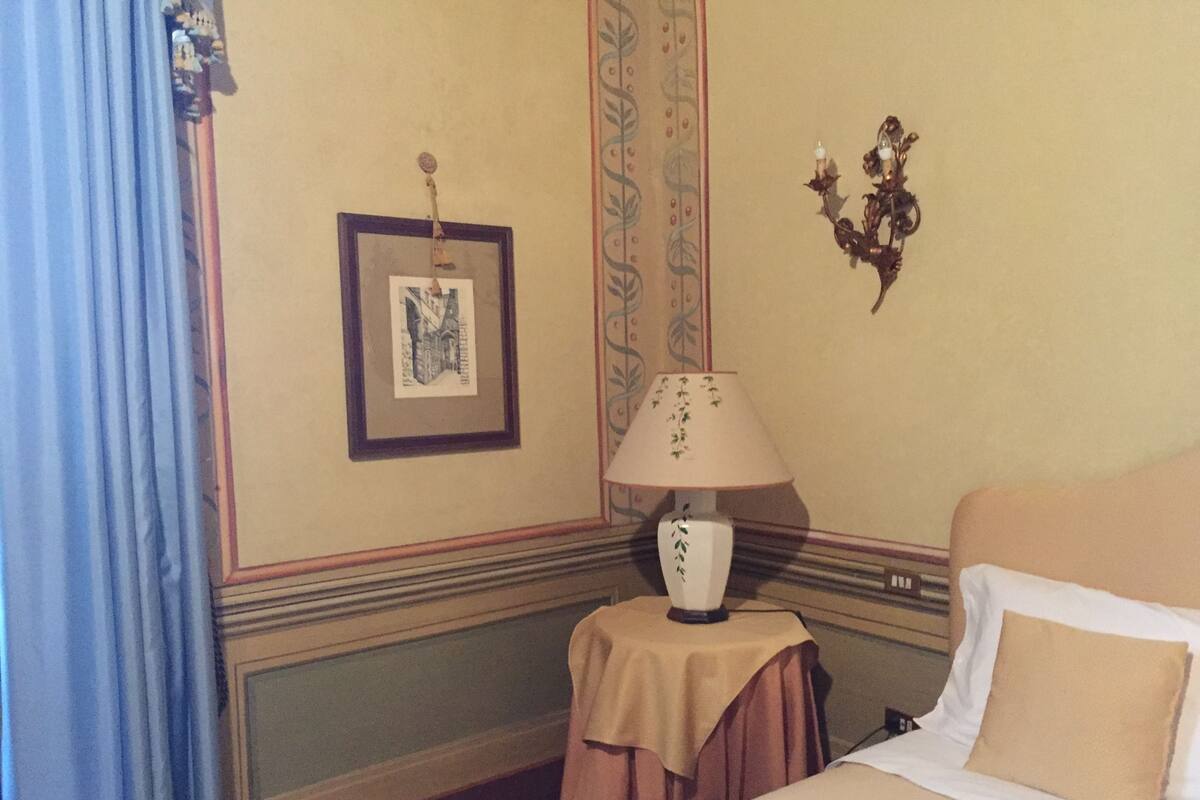 Hotellihuone, jossa on sänky, lamppuvarustettu pöytä, seinällä kehystetty taulu ja koristeellinen seinävalaisin.