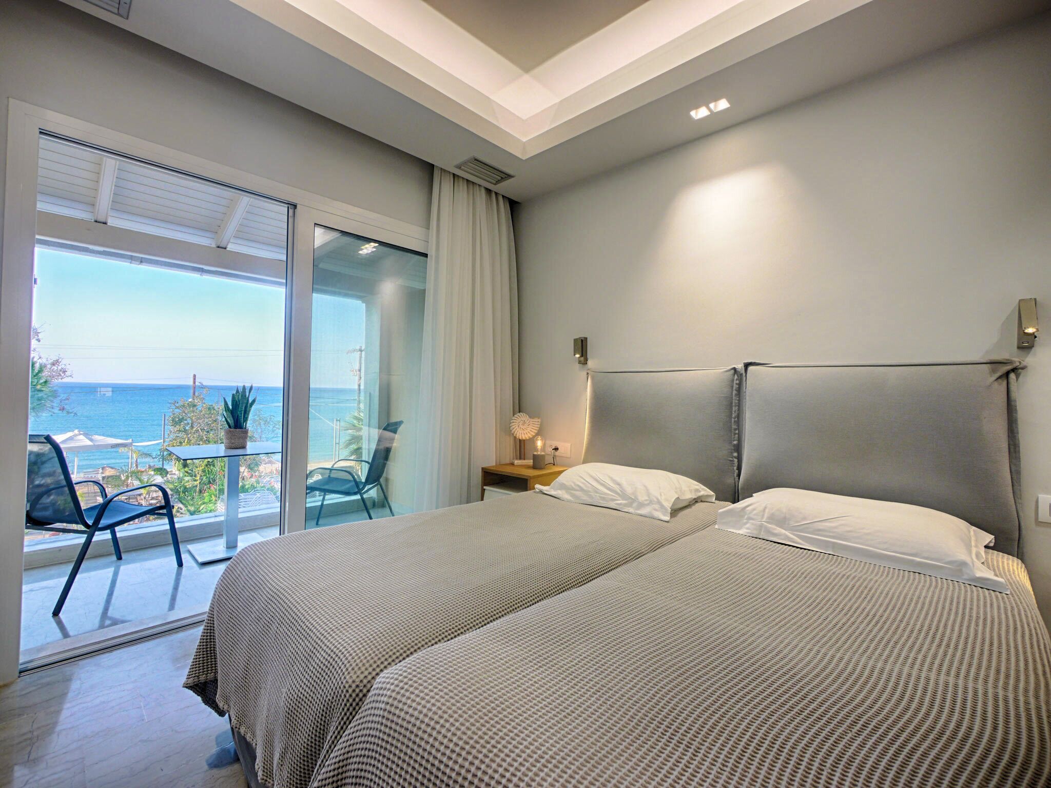 A bedroom with two beds, a balcony with a view of the sea, and a small table with a lamp.