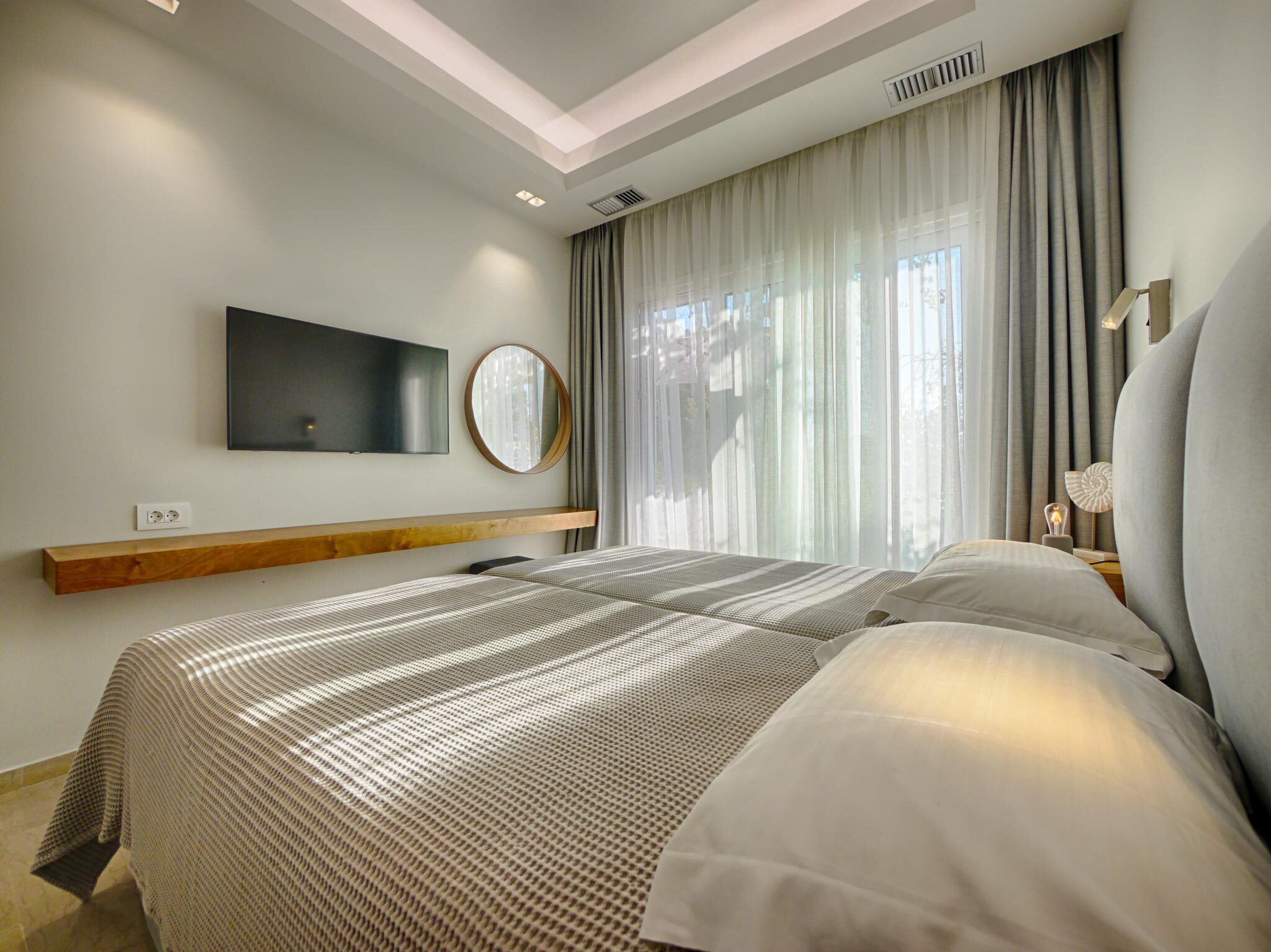 A modern hotel room with a large bed, a mounted TV, a round mirror, and a window with sheer curtains.
