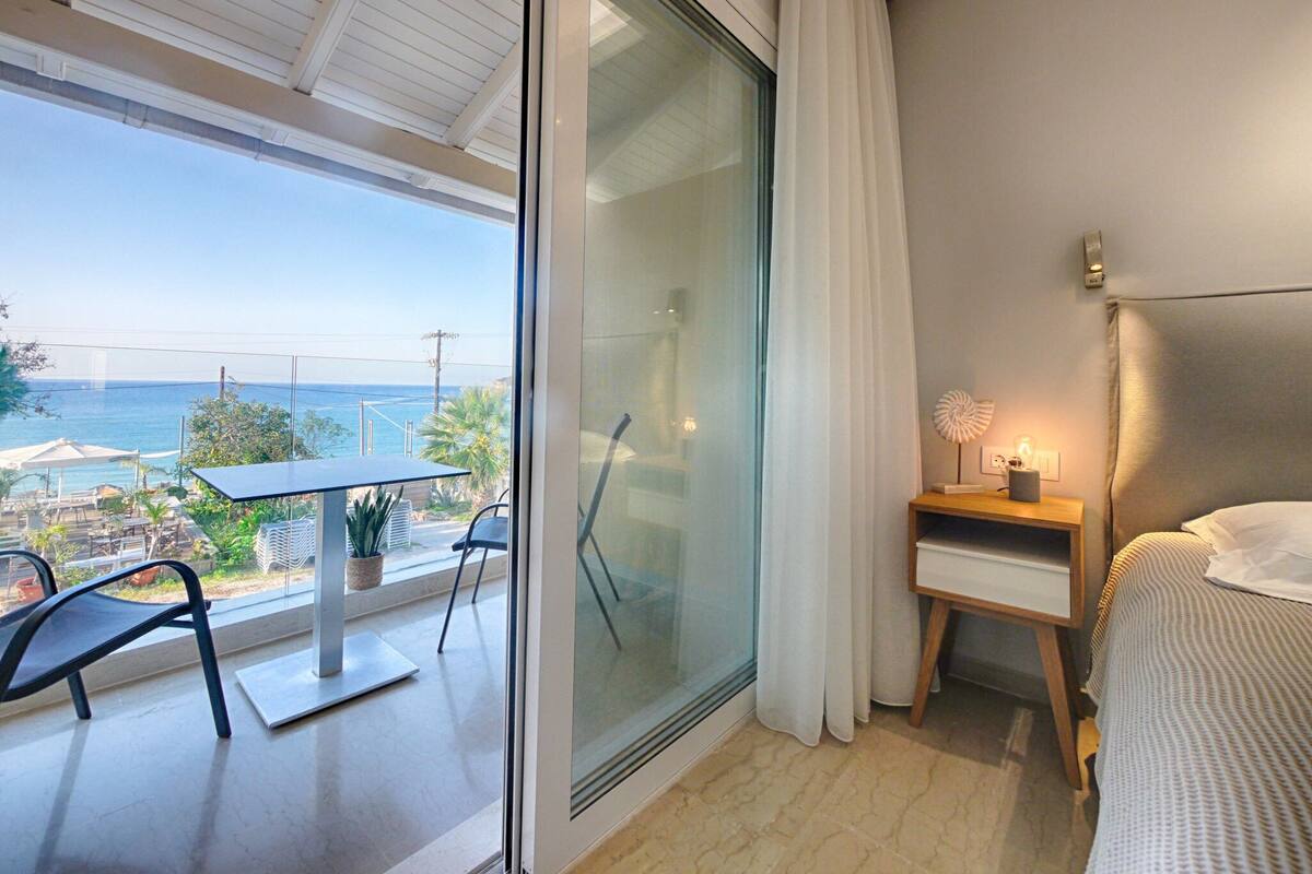 A balcony with a table and chairs, a bed with a headboard, a bedside table with a lamp, and a view of the sea and a bridge.