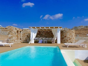 A pool area with a stone wall, a pergola with white curtains, and lounge chairs.