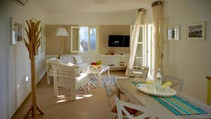 Una sala de estar bien iluminada con un sofá, una mesa de comedor con sillas, un televisor montado en la pared y un ventanal con cortinas.