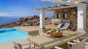 Zona junto a la piscina con mesa y sillas, pared de piedra y vista al mar.