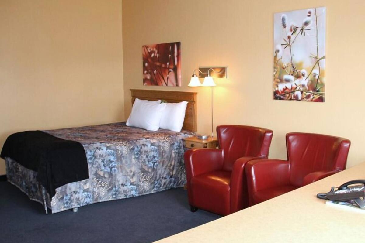Habitación de hotel con cama, dos sillas rojas, una mesita de noche, una lámpara y dos obras de arte en la pared.
