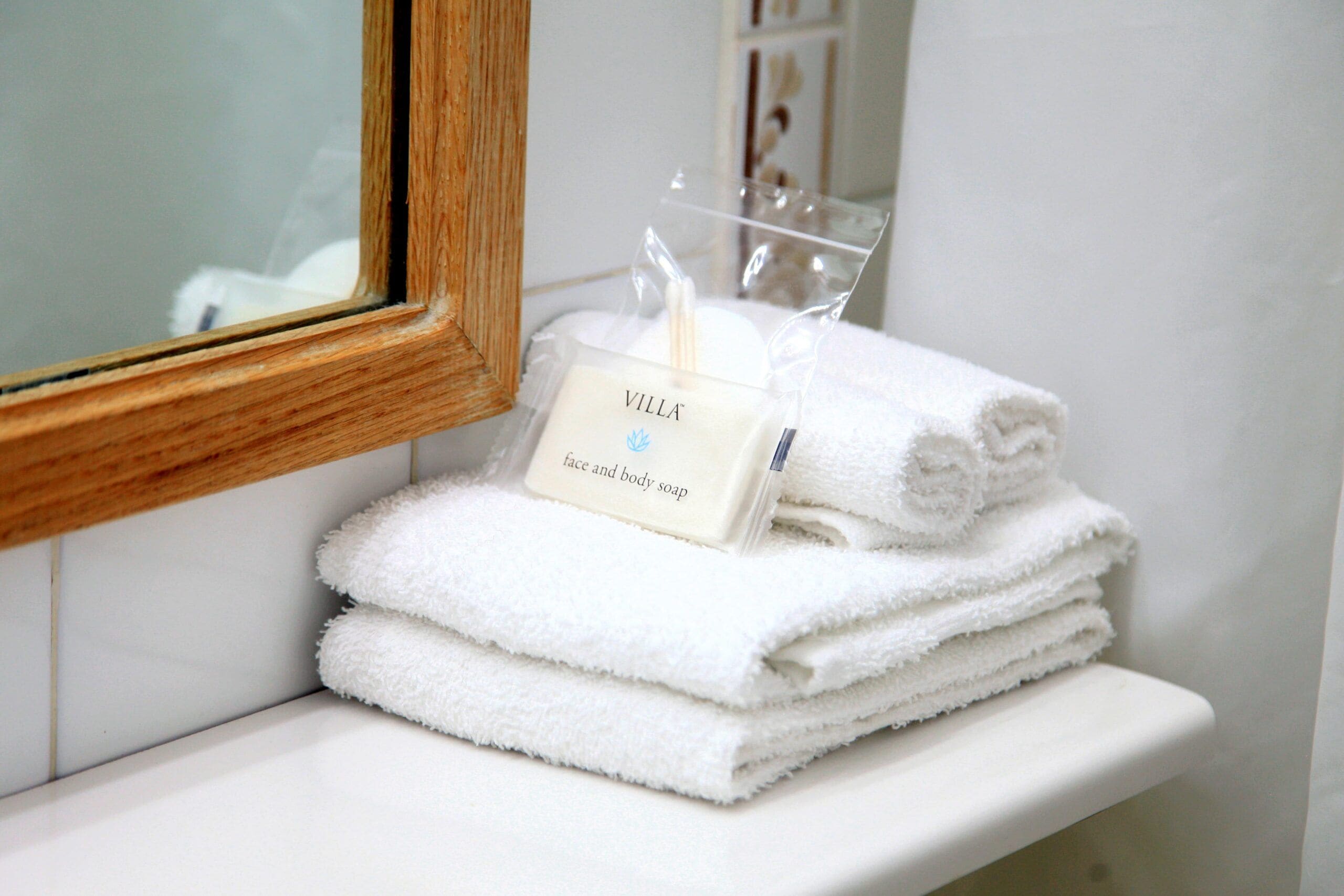 A stack of white towels on a white shelf with a wooden-framed mirror above them. A small white box with the text 'VILLA face and body soap' is placed on the towels.