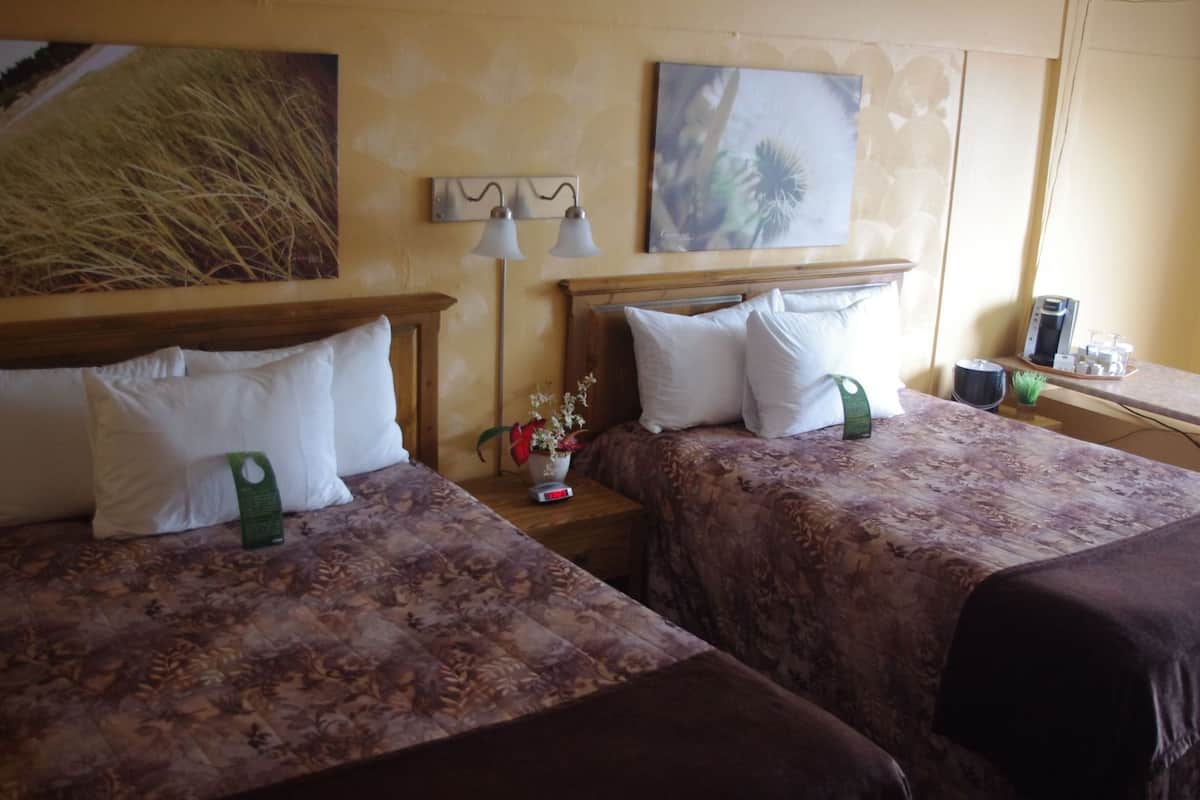 Habitación de hotel con dos camas, una mesita de noche con lámpara, cafetera y un cuadro en la pared.
