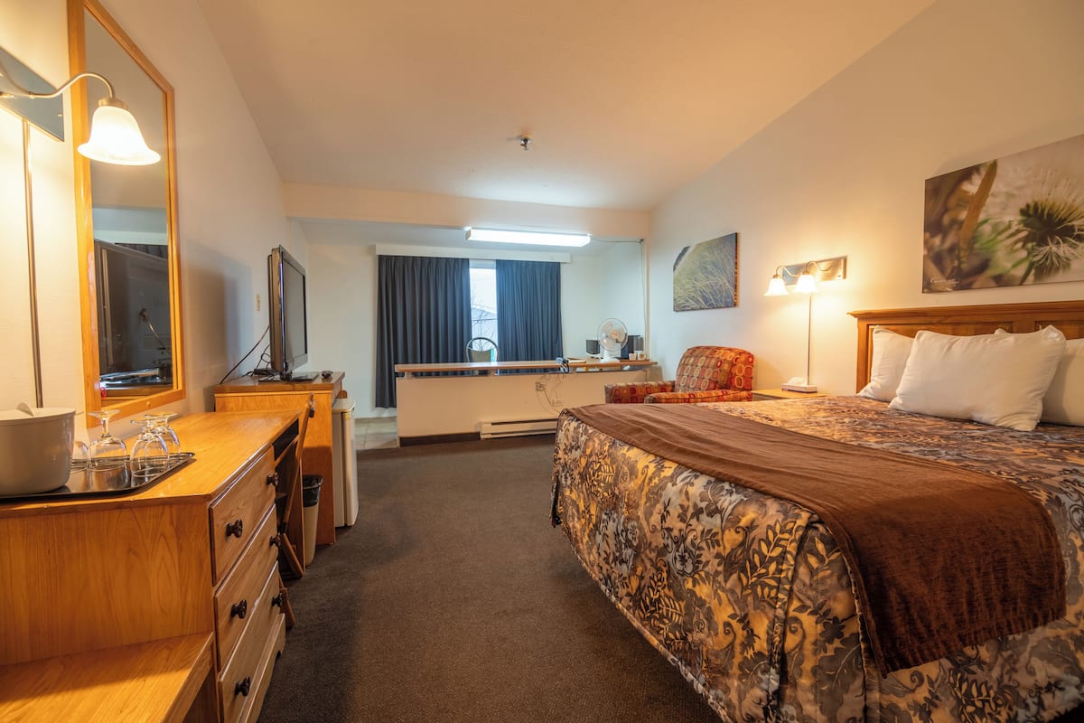 Habitación de hotel con una cama grande, un aparador de madera, un escritorio y una silla. Se observa una ventana con cortinas, un espejo y un cuadro en la pared.