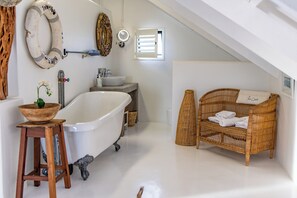 A bathroom with a freestanding bathtub, a wooden stool with a bowl, a wicker chair, and a small table with a flower vase. - SaWestCoast & Beyond (Paternoster)
