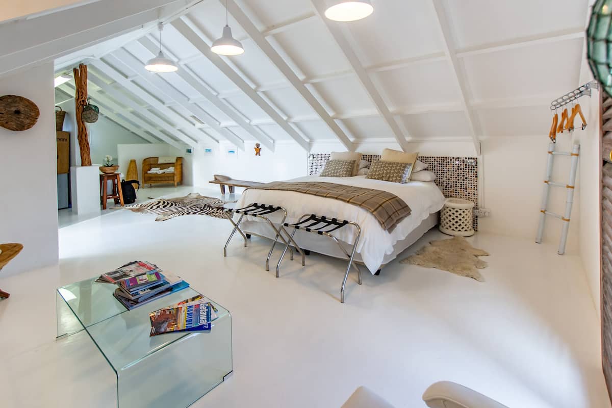A modern bedroom with a loft-style ceiling, a large bed with multiple pillows, a glass coffee table with magazines, and a rustic rug.