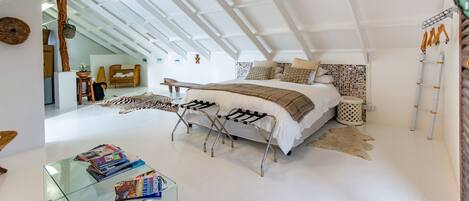 Un dormitorio moderno con techo estilo loft, una cama grande con muchas almohadas, una mesa de centro de vidrio con revistas y una alfombra rústica.