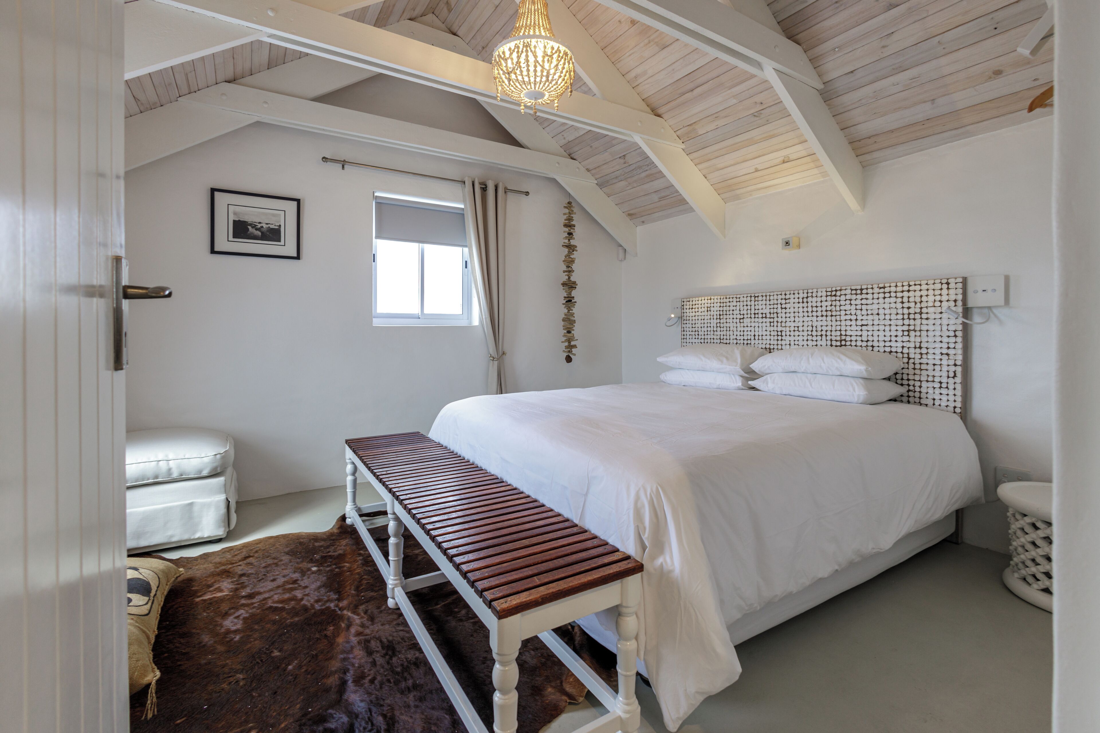 A bedroom with a wooden bench, a bed with a white headboard, a window with curtains, and a chandelier.