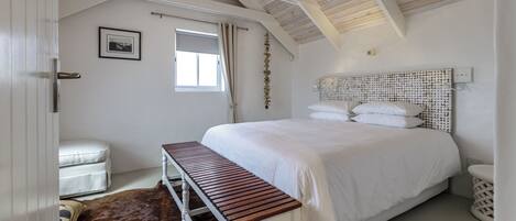 A bedroom with a wooden bench, a bed with a white headboard, a window with curtains, and a chandelier.