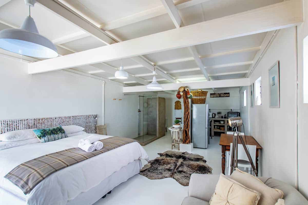 A spacious bedroom with a large bed, a desk, and a chair. There is a glass shower area and a kitchen visible in the background.