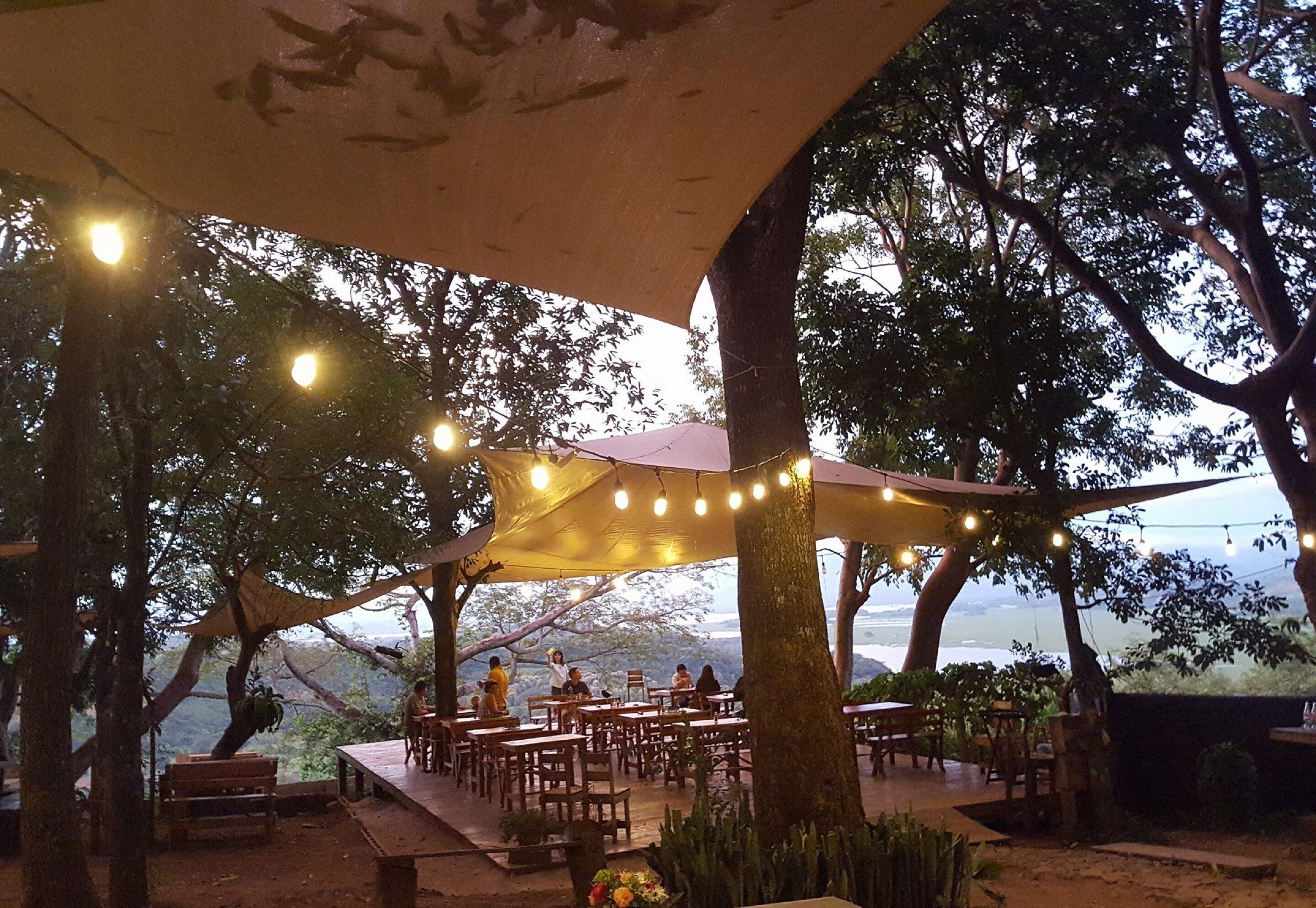 An outdoor dining area with tables and chairs under a canopy, surrounded by trees and overlooking a scenic view.