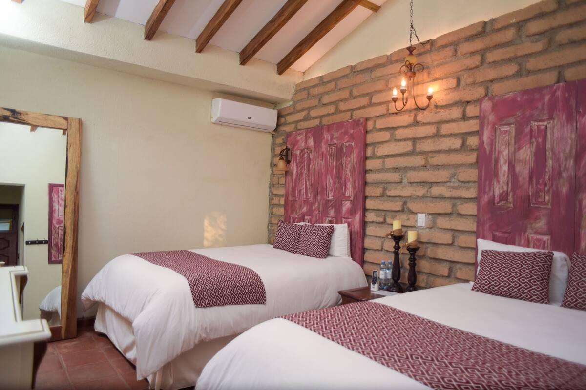 A hotel room with two beds, a brick wall, wooden beams, a hanging light fixture, and a mirror.