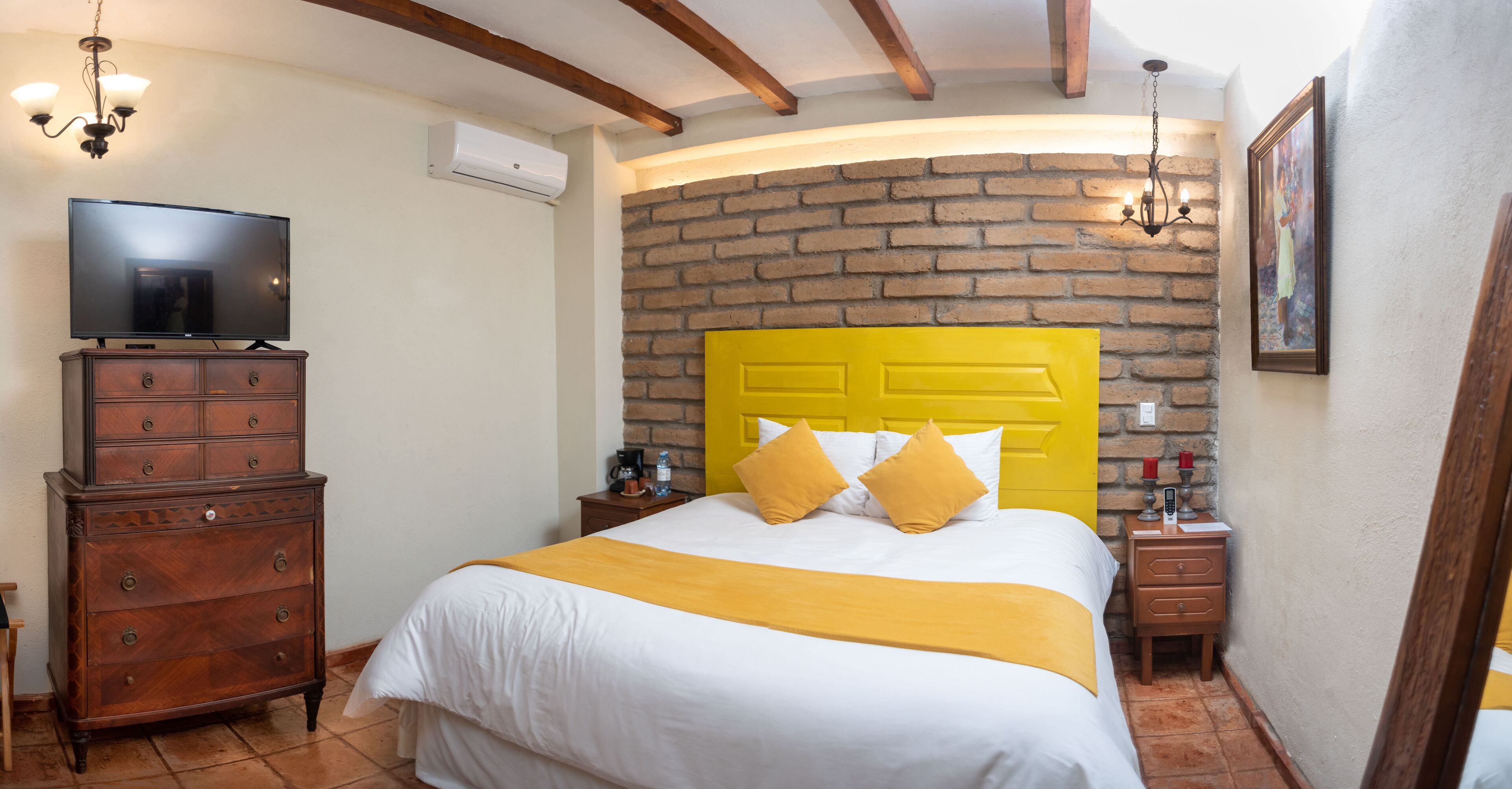 Una habitación de hotel con pared de ladrillo, cabecera amarilla, televisor de pantalla plana, muebles de madera y candelabro.