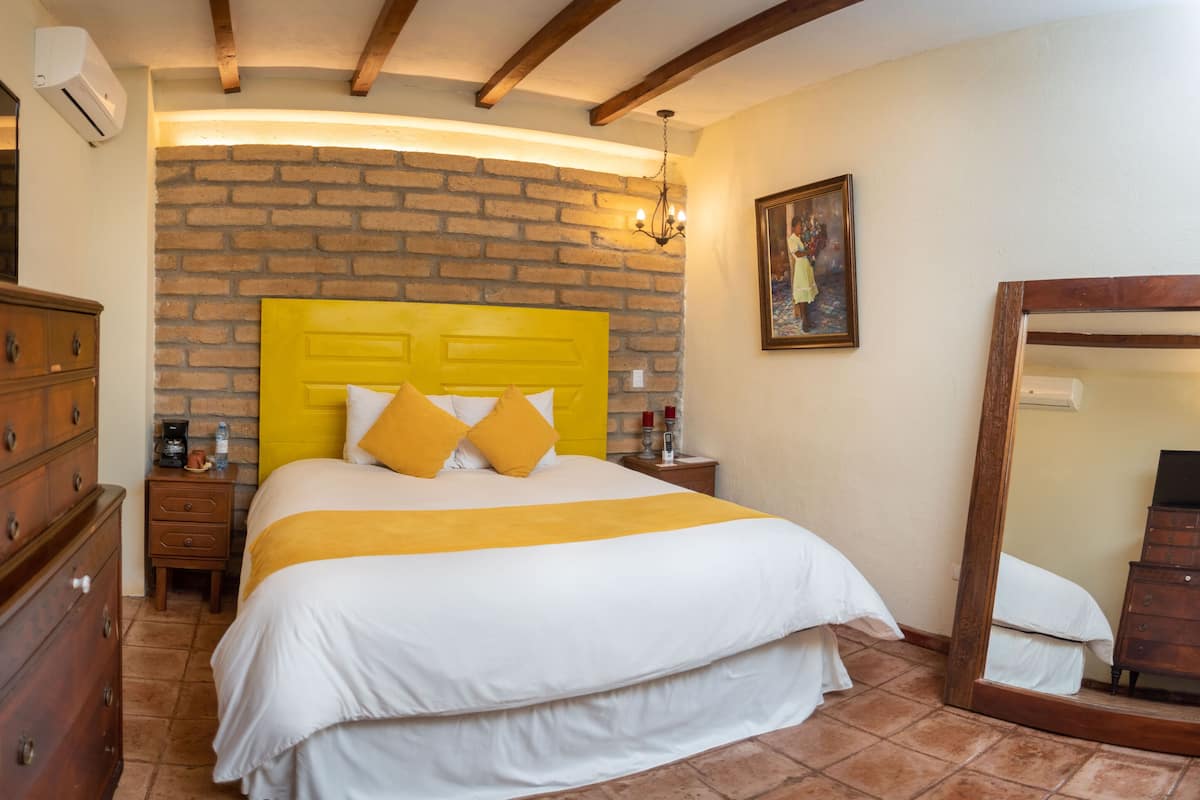 A bedroom with a brick wall, a wooden headboard, a bed with white and yellow bedding, a wooden dresser, a nightstand with a lamp, and a mirror.