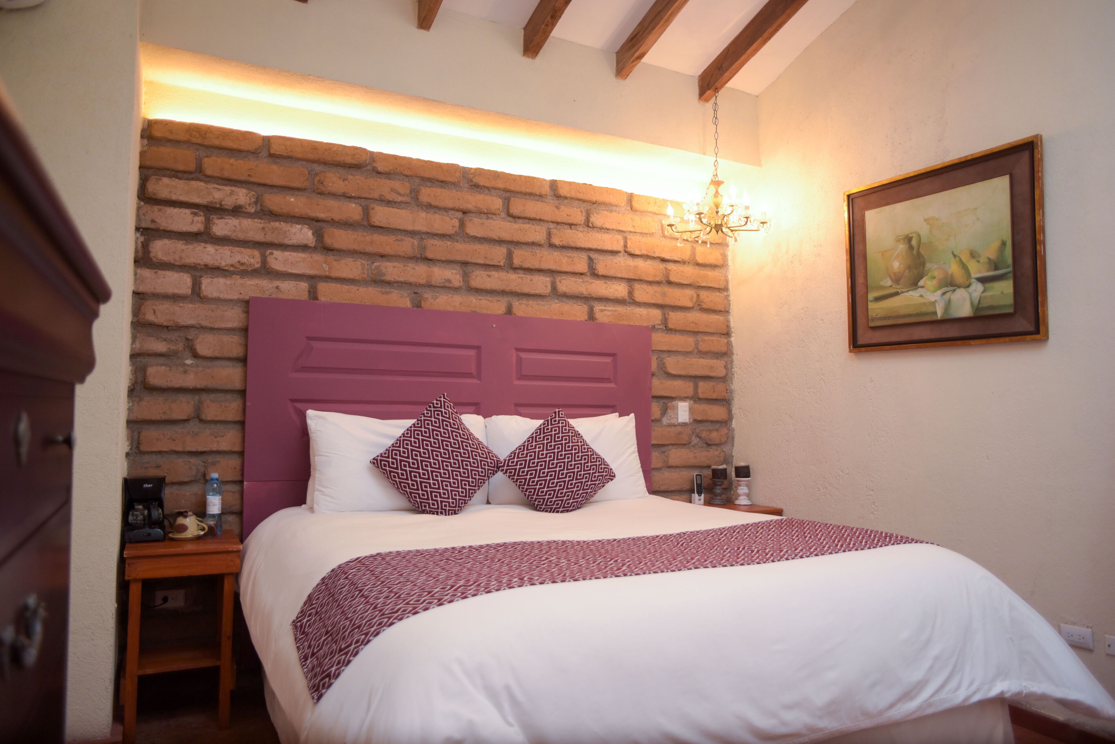 Quarto com parede de tijolos, cabeceira de madeira, cama branca com almofadas estampadas, mesa de cabeceira com garrafa de água e um quadro na parede.