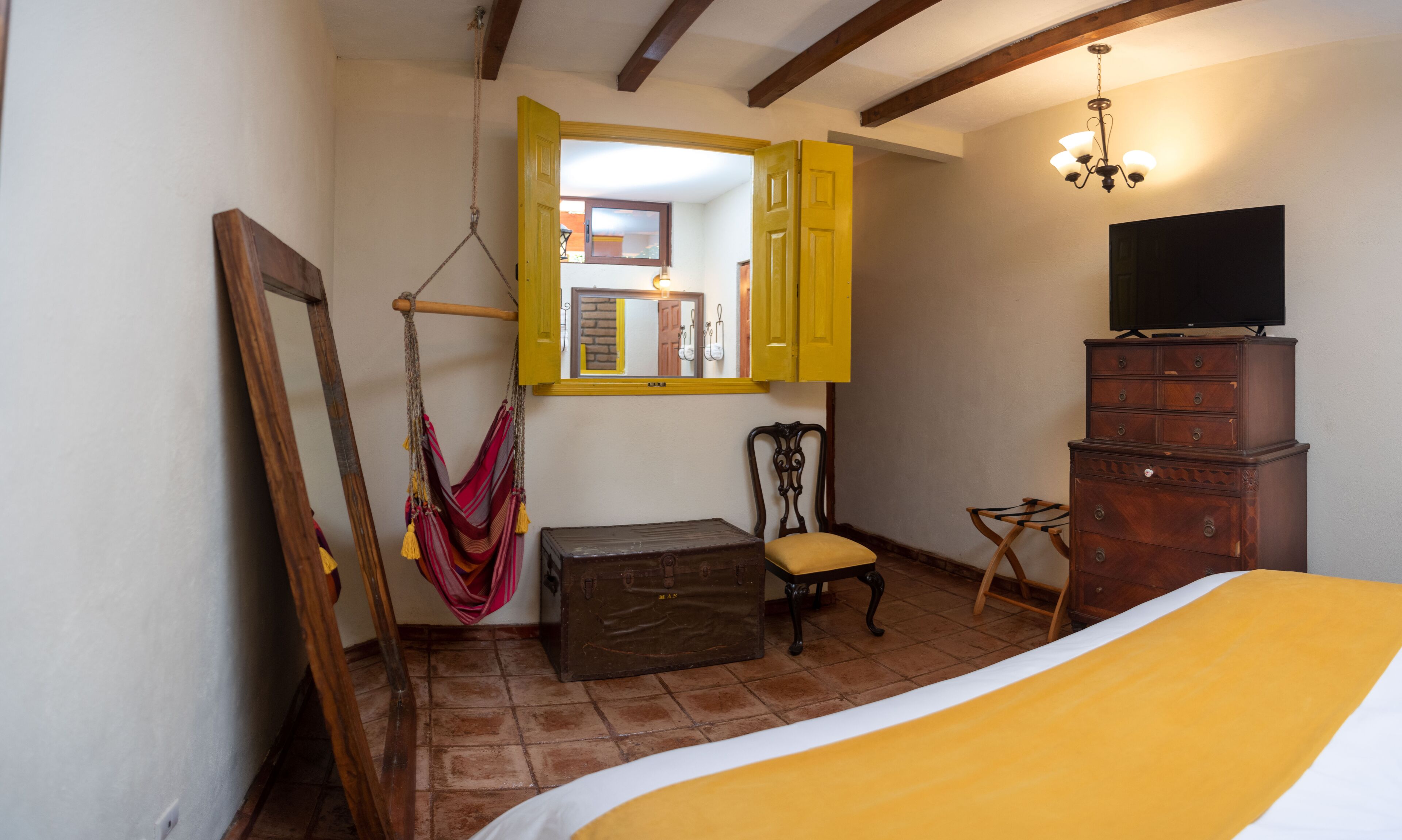 A bedroom with a bed, a wooden dresser, a chair, a hanging hammock, and a television on a dresser.