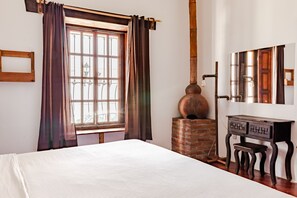 Une pièce comprenant un lit, une armoire en bois, un poêle en briques et une fenêtre avec des rideaux.
