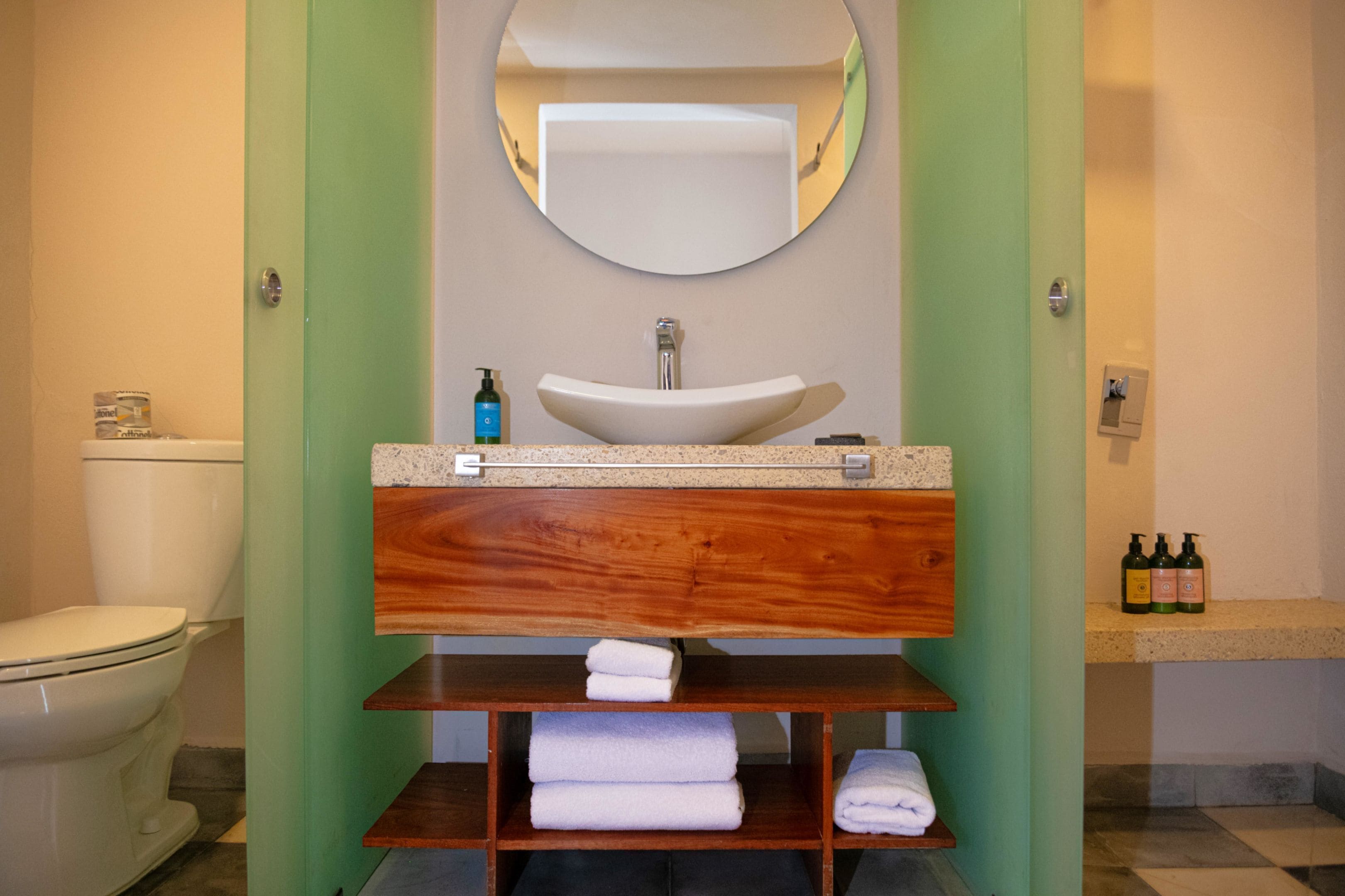 A bathroom with a white sink, a mirror above it, and a toilet to the left. There are shelves with folded towels and bottles of toiletries.