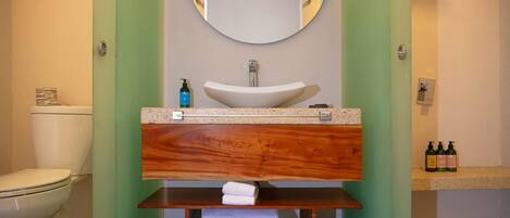 A bathroom with a white sink, a mirror above it, and a toilet to the left. There are shelves with folded towels and bottles of toiletries.