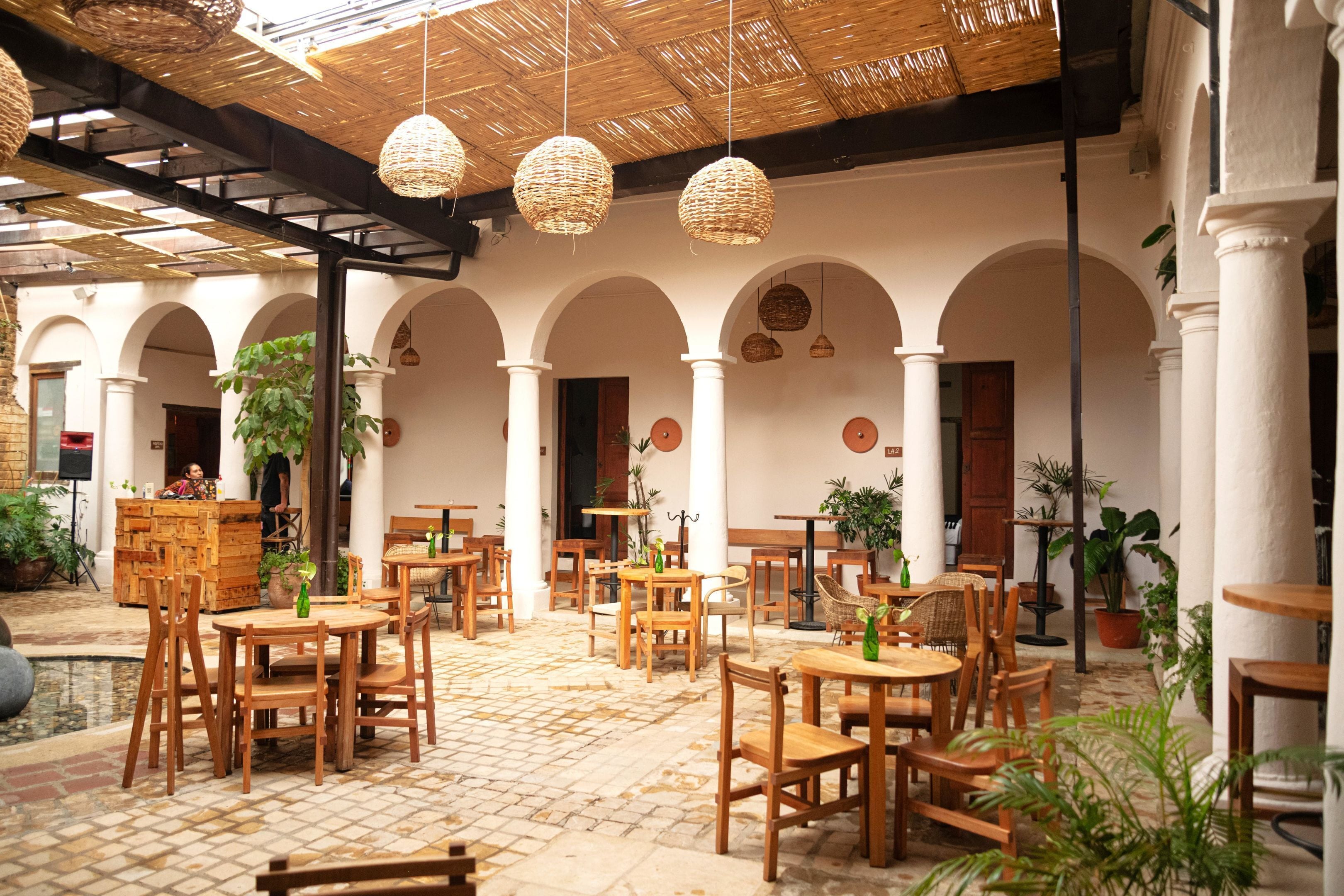 Un patio interior con mesas y sillas de madera, suelo de baldosas y lámparas de mimbre colgantes.
