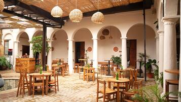 Un patio interior con mesas y sillas de madera, suelo de baldosas y lámparas de mimbre colgantes.