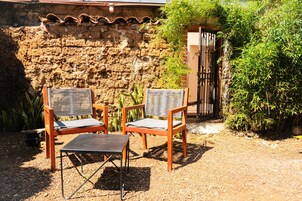 Deux chaises en osier et une petite table sont disposées dans un coin du jardin, adossées à un mur de pierres rustiques et à une porte en métal.