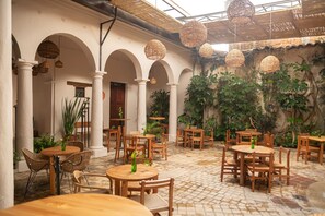 Une cour avec des tables et des chaises rondes, entourée d'arcades et de colonnes, agrémentée de lampes en osier suspendues et de plantes en pot.