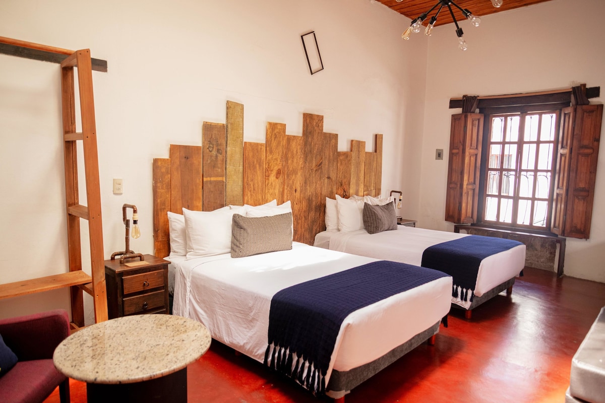 Quarto de hotel com duas camas, cabeceira de madeira, uma pequena mesa e uma janela com venezianas de madeira.