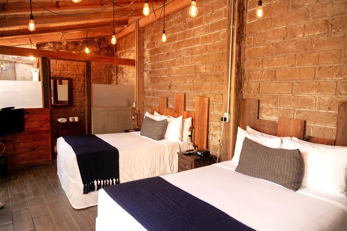 Quarto de hotel com duas camas, cabeceiras de madeira e paredes de tijolo aparente.
