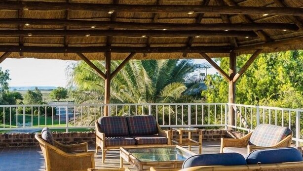 Un área de estar al aire libre techada con mobiliario de mimbre y una mesa de vidrio, con vistas a un paisaje exuberante con palmeras y una vista distante del mar.