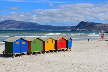 A row of colorful beach huts on a sandy beach with a mountain in the background.
