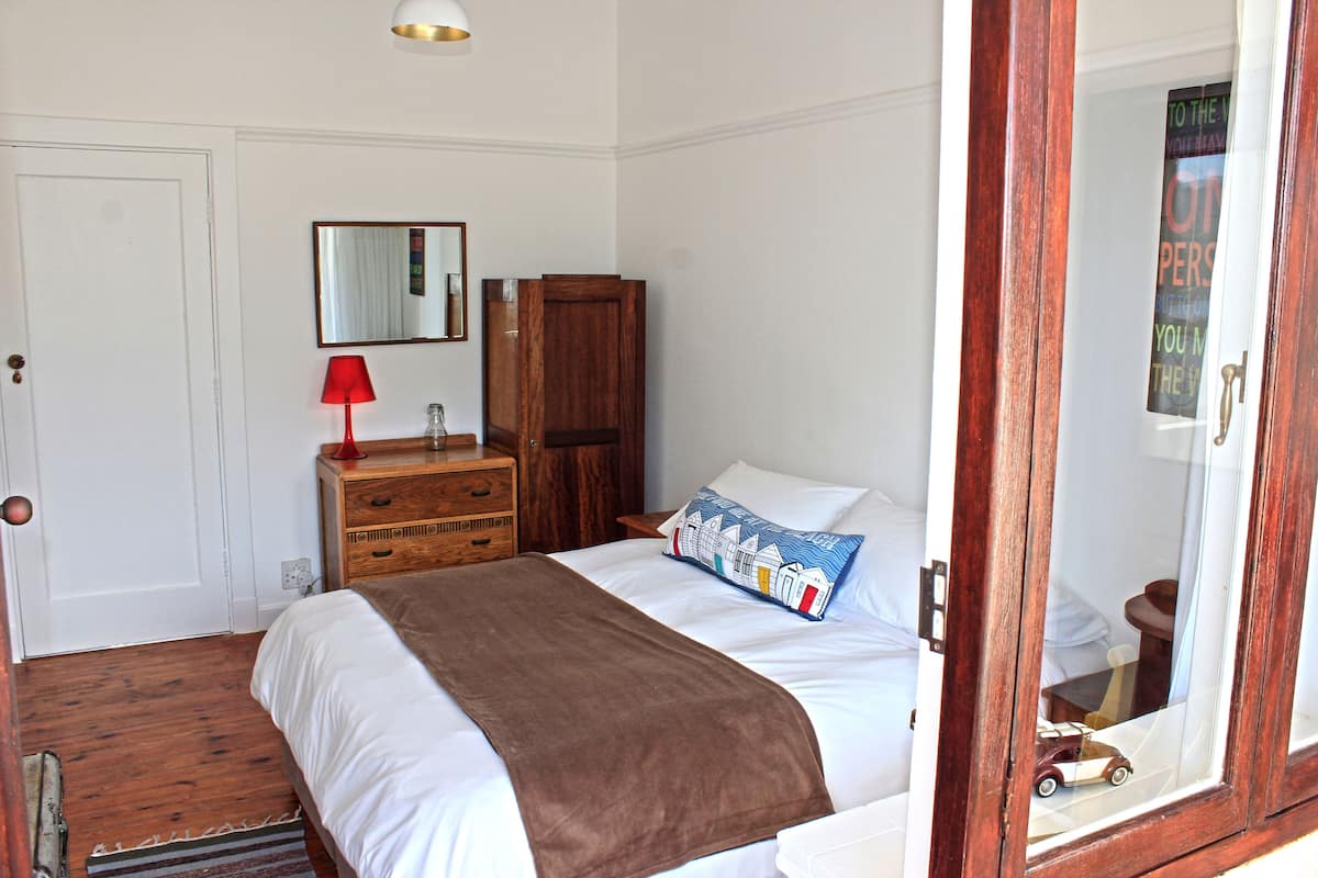 A bedroom with a bed, a wooden dresser, a red lamp, a mirror, and a window with a view of a building.