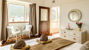 Une chambre d’hôtel avec deux fauteuils en osier blancs, un lit avec des serviettes marron, une commode blanche avec un miroir et une vue sur un paysage montagneux par la fenêtre.